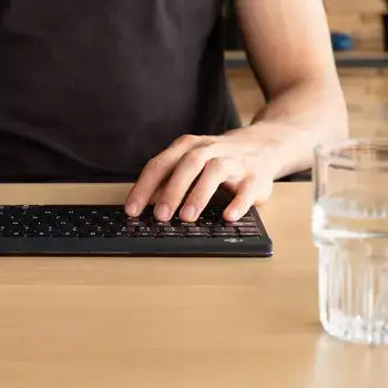 Person typing on Ergo-R Black mini Bluetooth keyboard, showcasing ergonomic design on a wooden desk with a glass of water nearby.